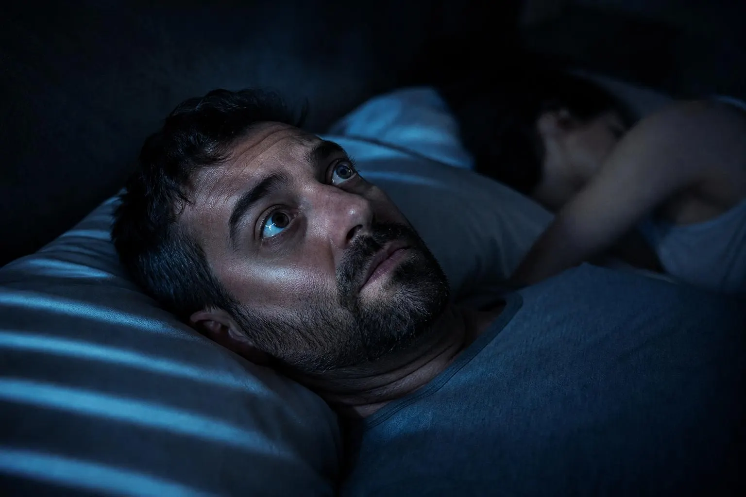 Man lying awake in bed at 3 AM staring at the ceiling, partner asleep beside him, blue moonlight across his face